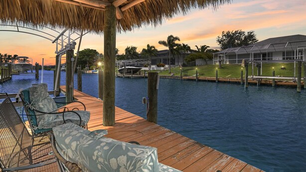 A wooden deck with lounge chairs overlooking a body of water, a thatched roof structure, and a building in the background. - Fully stocked & Pet Friendly! Bikes, pool table, hot tub & more! Villa Aleena - SWFL Rentals (Cape Coral)