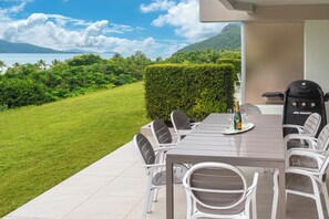 An outdoor dining area with a view of the ocean and hills, a grill, and a table set with chairs and a bottle.