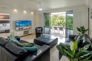A modern living room with a flat-screen TV, black leather sofas, a dining area with a table and chairs, and a sliding glass door leading to a balcony with a view of palm trees.