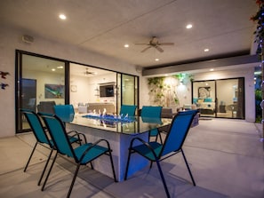 A modern dining area with a glass table and blue chairs, a ceiling fan, and a view into the adjacent living space. - Casa Azul - Luxury 4BR/4BA Private Compound (Palm Springs)