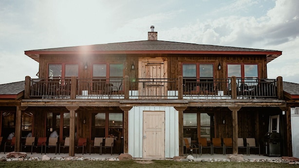 A two-story wooden house with a balcony, large windows, and a covered patio area.