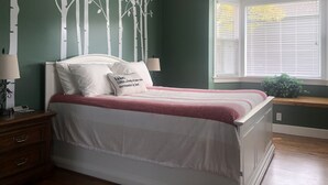 A bedroom with a bed, a nightstand, a lamp, a window with blinds, and a wall with a tree design.