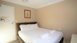 A neatly made bed with white linens and two rolled towels, a wooden headboard, and a framed picture on the wall.