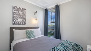 A bedroom with a bed, a headboard, a wall-mounted light, and a view of a balcony. - The Waves - Sit Back And Watch The Waves Roll In (Port Fairy)
