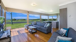 A spacious living room with a large window offering a view of the outdoors, a grey sofa, a coffee table, and a flat-screen TV. - The Waves - Sit Back And Watch The Waves Roll In (Port Fairy)