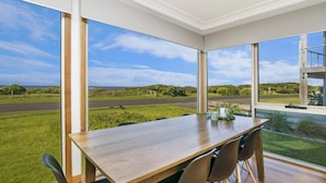 A dining area with a large table and chairs, large windows offering a view of a road and greenery. - The Waves - Sit Back And Watch The Waves Roll In (Port Fairy)