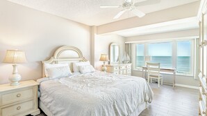 Ein Schlafzimmer mit einem großen Bett, einem Nachttisch mit Lampe, einer Kommode, einem Sessel und Meerblick durch große Fenster.