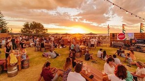 Een druk evenement buiten, met mensen die op vaten en tafels zitten, onder een zonsondergang.