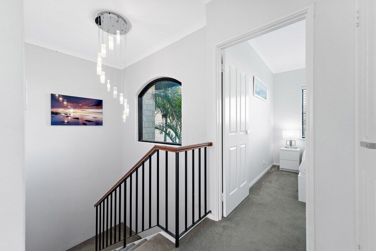Un pasillo con una escalera, paredes blancas, un cuadro enmarcado, una ventana con vistas y una mesita de noche con una lámpara.