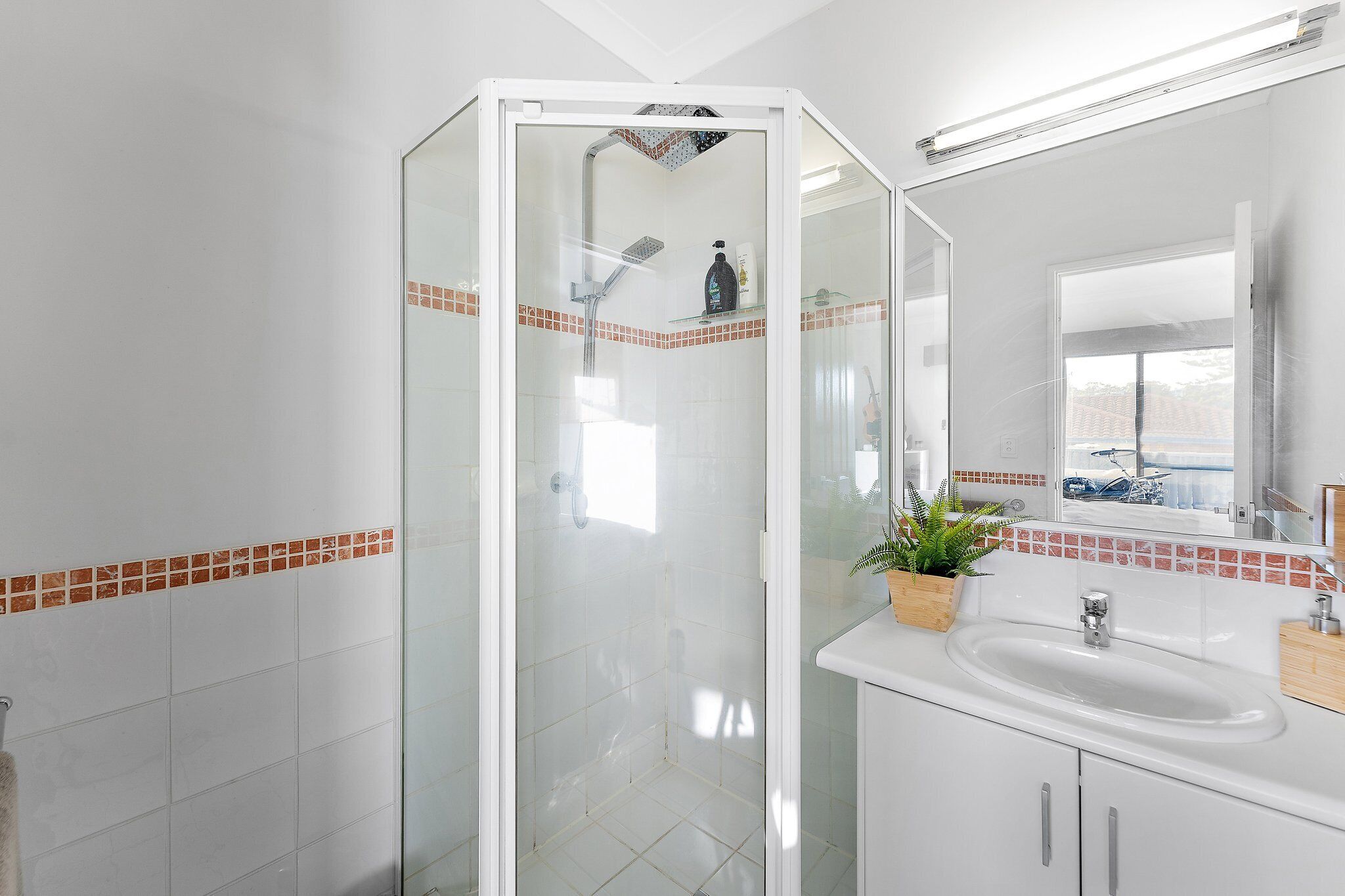 Une salle de bain moderne avec une douche à l’italienne en verre, des carreaux blancs, un lavabo avec un miroir et une fenêtre donnant sur un paysage.