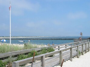 A wooden boardwalk leading to a beach with a flagpole, a lifeguard tower, and a clear view of the ocean.