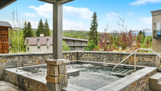 A stone hot tub with a metal ladder, surrounded by a stone wall, set against a backdrop of buildings and trees.
