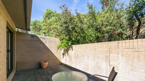Une terrasse avec une table ronde en verre et deux chaises, un mur en briques et un pot de fleurs.