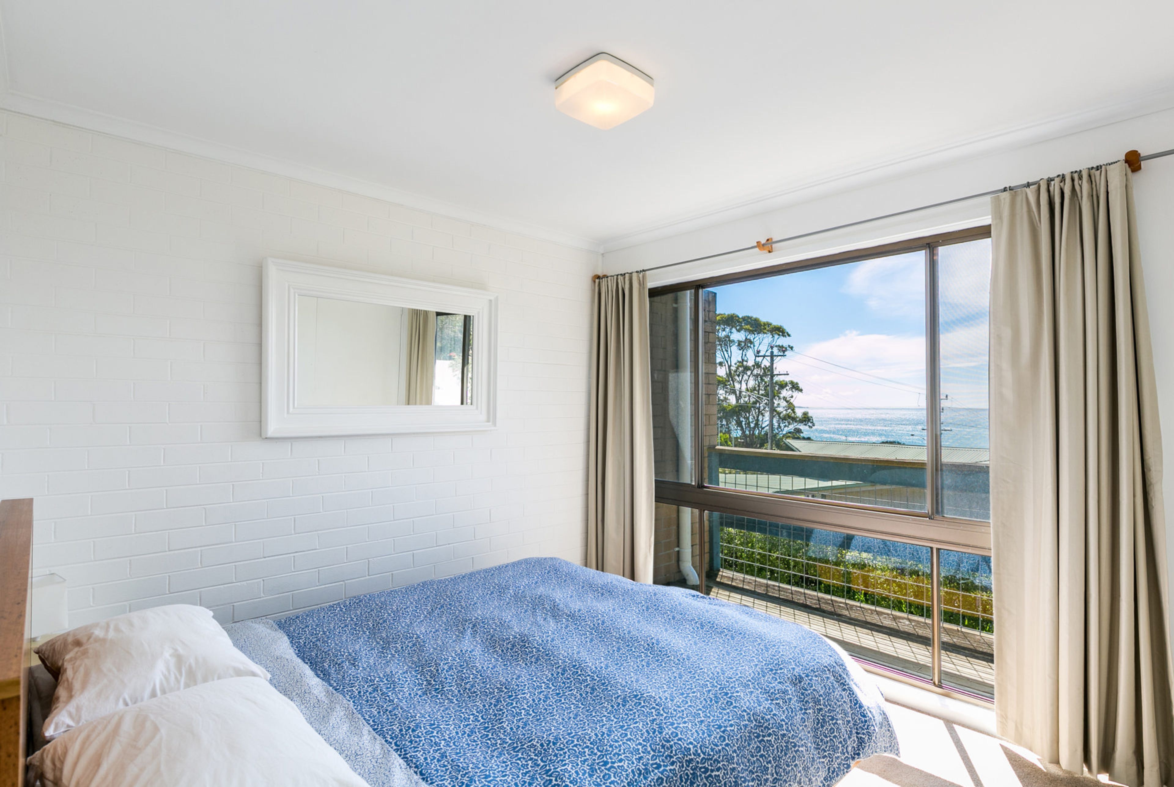 A bedroom with a bed, a window with curtains, a mirror on the wall, and a view of the outdoors.