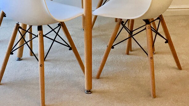 Ein runder, weißer Tisch mit vier weißen Stühlen, jeweils mit Holzbeinen und schwarzen Metallstreben. Auf dem Tisch steht eine transparente Glasvase mit weißen Blumen.