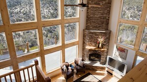 A spacious living room with large windows, a stone fireplace, and a TV. - Rock Canyon Ranch Lodge 2 Zion National Park (Orderville)