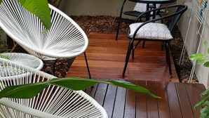 Una terraza de madera con sillas de mimbre blanco y una mesita, rodeada de exuberante follaje verde.