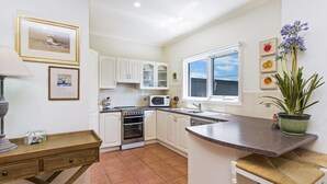 Una cocina compacta con gabinetes blancos, un microondas, un horno y un lavamanos. Hay una ventana que proporciona luz natural y una pequeña mesa con una lámpara.