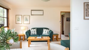 Un salon avec un canapé vert, une table basse en bois, une lampe, des reproductions botaniques encadrées accrochées au mur et un climatiseur fixé au plafond.