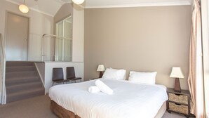 Une chambre d’hôtel avec un grand lit, deux lampes de chevet, une petite table avec une lampe, une chaise, un escalier et une vue sur l’extérieur.