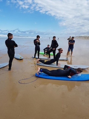 Un groupe de personnes se trouve sur une plage, certaines tenant des planches de surf, d’autres debout et d’autres allongées sur le sable.
