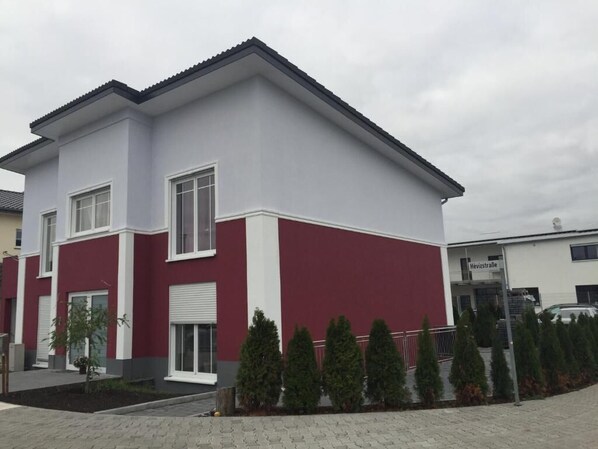 A two-story building with a red and white facade, a visible garage door, and a small garden with coniferous trees. - Appartements Funk (Pfungstadt)