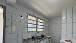A kitchen with white tiles, a window with blinds, a sink, and a countertop. - Encanto na Praia da Daniela \- Florianópolis \- SC (Florianópolis)