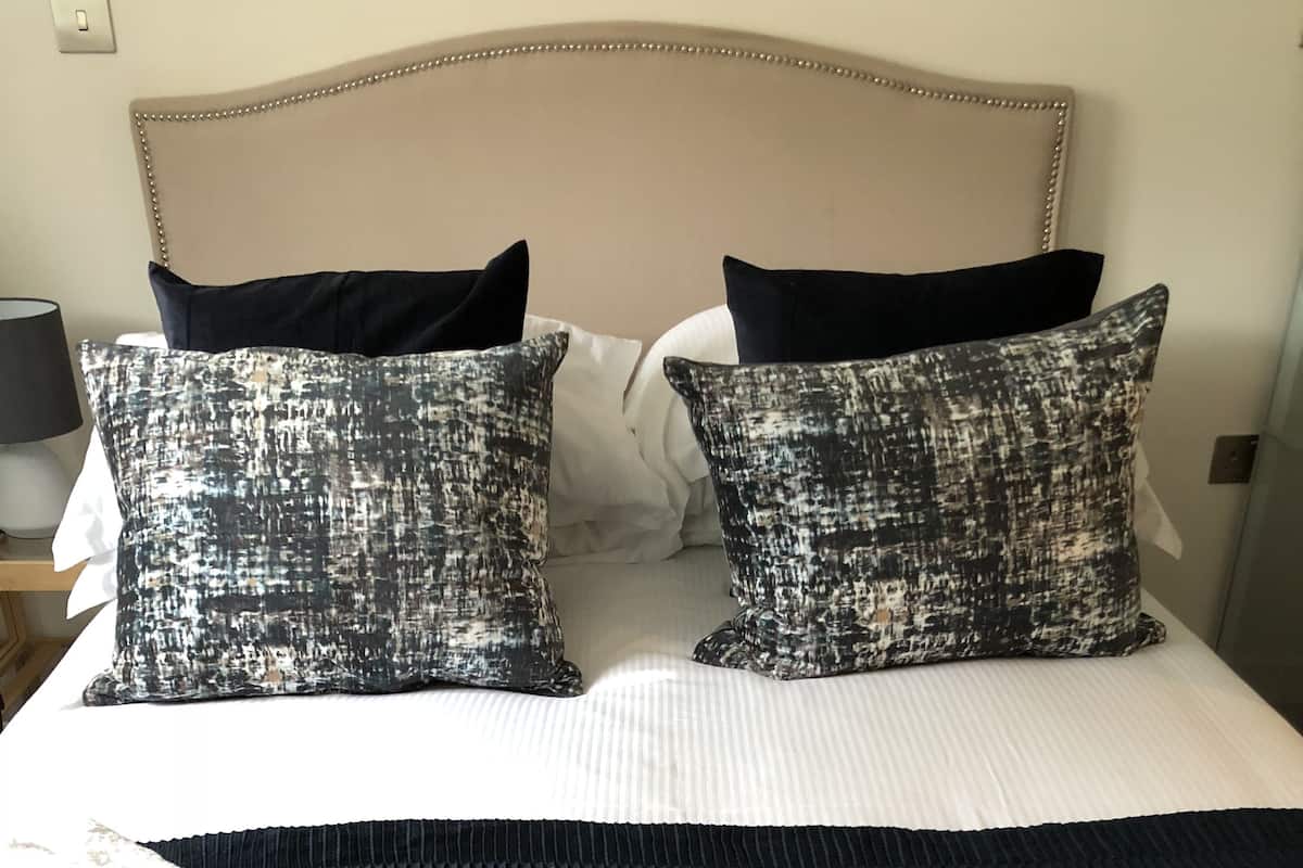 Cama bem arrumada com edredom branco, duas almofadas estampadas, manta escura e toalhas dobradas.
