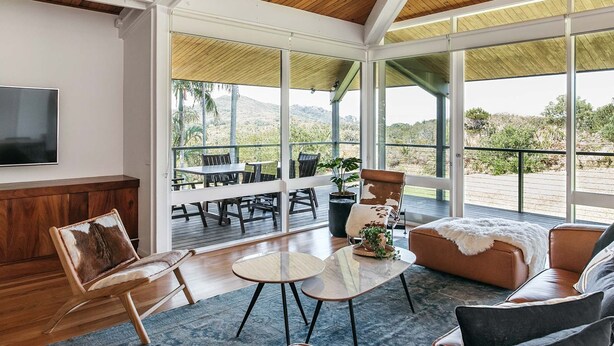 A spacious living area with a large window overlooking a scenic landscape, a comfortable seating arrangement, and a flat-screen TV mounted on the wall. - Cape Breeze - 5 BR home with headland views (Byron Bay)