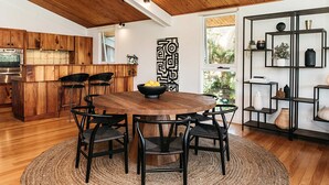 A modern kitchen with wooden cabinetry, a central island, and a dining area with a round table and chairs. - Cape Breeze - 5 BR home with headland views (Byron Bay)
