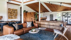 A spacious living area with a high ceiling, wooden beams, a brown leather sofa, a round coffee table, and a dining area with a wooden table and chairs. - Cape Breeze - 5 BR home with headland views (Byron Bay)