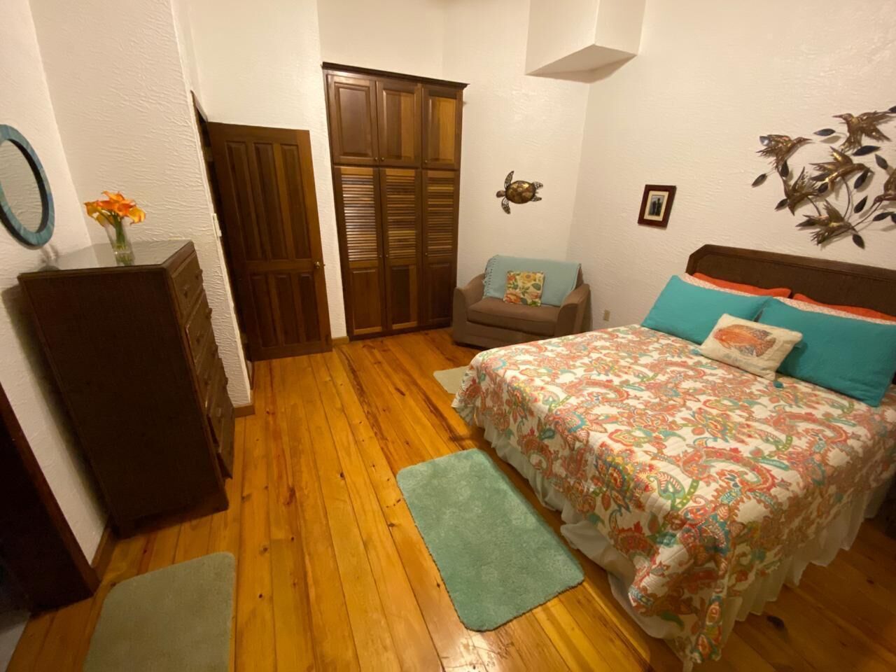 Quarto com piso de madeira, uma cama com roupa de cama floral, uma cadeira, um guarda-roupa e uma pequena mesa com um vaso de flores.