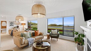 A spacious living room with a large window offering a view of a road and greenery. There are two sofas, a coffee table, and a TV mounted on the wall. - 10 Ocean Drive - one of those properties that  will have you never wanting to leave (Port Fairy)
