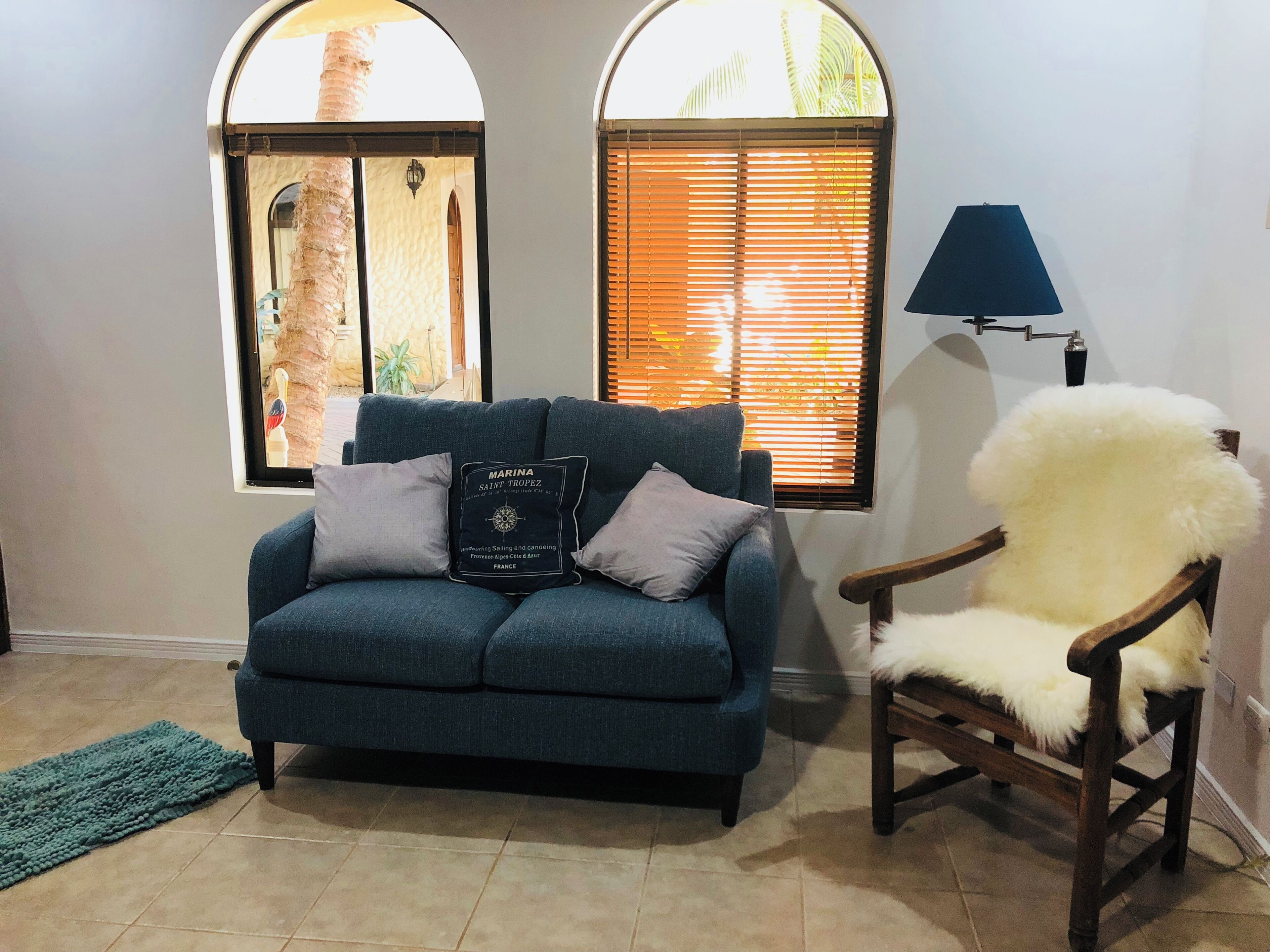 Un salón con un sofá azul, una silla de madera, una lámpara y dos ventanas con persianas.