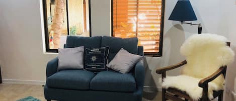Un salón con un sofá azul, una silla de madera, una lámpara y dos ventanas con persianas.