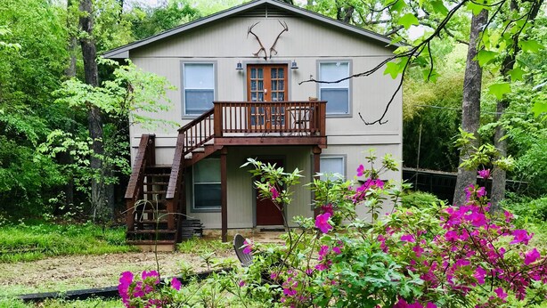 A two-story house with a balcony, surrounded by lush greenery and vibrant pink flowers.