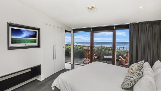 A modern bedroom with a large bed, a TV on the wall, and a balcony with ocean views. - Hearns Beachside Villa 1 (Port Fairy)