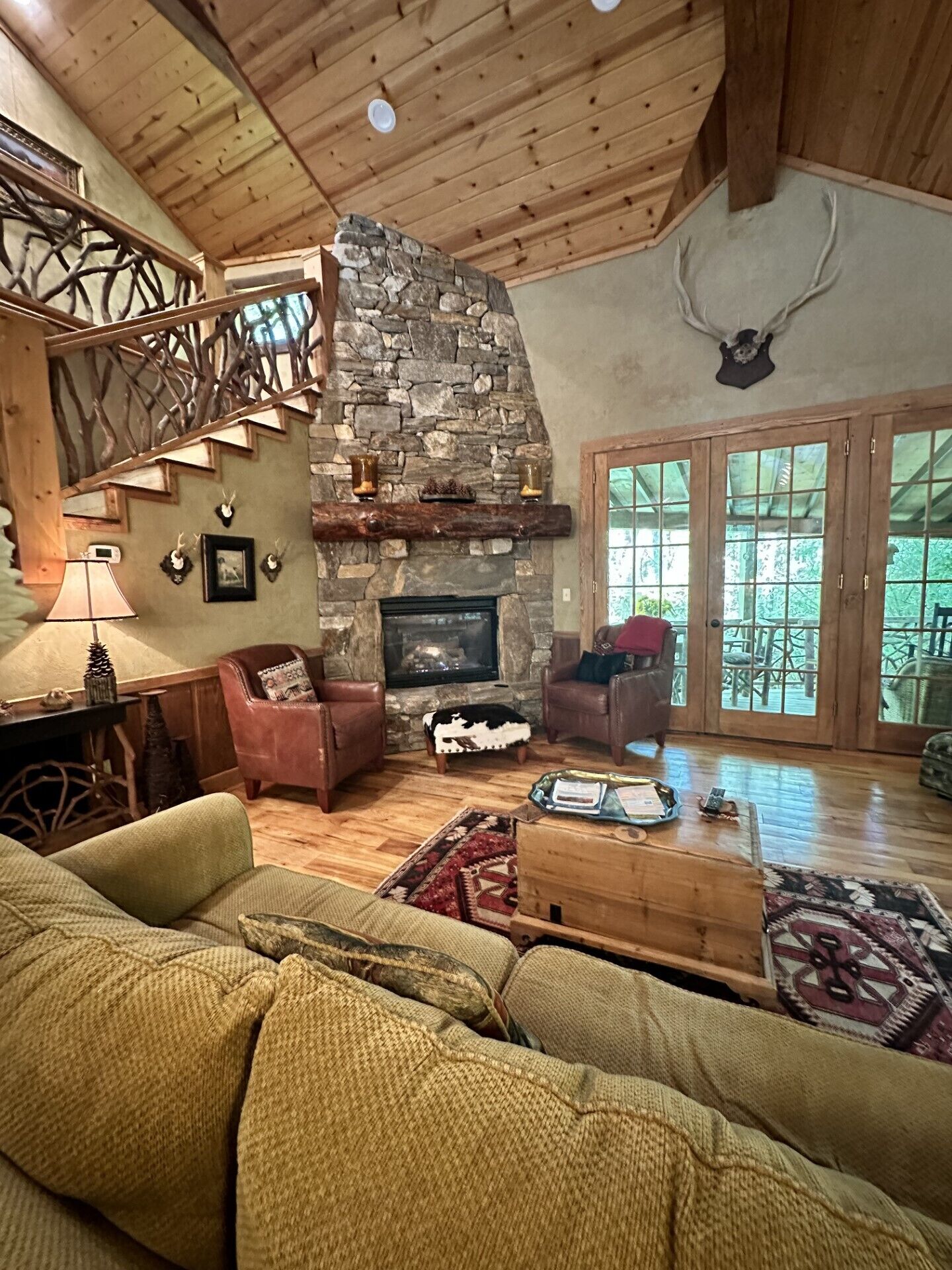 A cozy living room with a stone fireplace, wooden staircase, leather sofas, and a large window with a view of the outdoors.