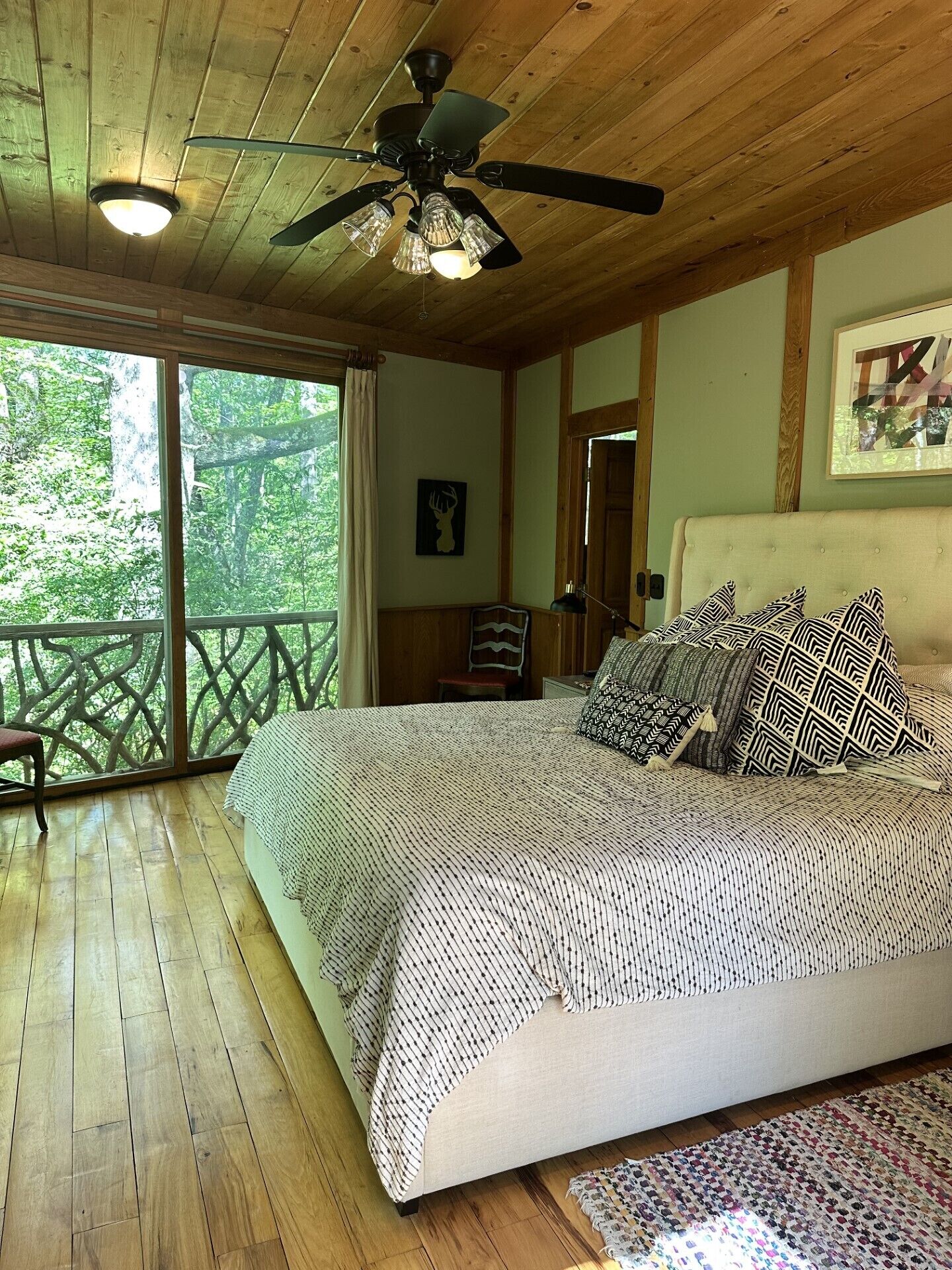 A bedroom with a bed, a ceiling fan, a window with a view of trees, and a wooden floor.