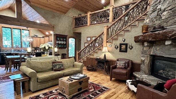A spacious living area with a high ceiling, wooden beams, a stone fireplace, a staircase with a wooden railing, and a kitchen in the background. - Hidden Hollow 2 BR 2 baths WALK to town (Highlands)