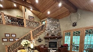 A spacious living room with a stone fireplace, wooden flooring, a staircase with a wooden railing, and a large window offering a view of the outdoors. - Hidden Hollow 2 BR 2 baths WALK to town (Highlands)