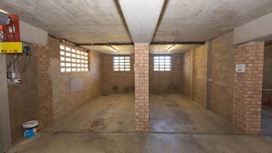 Ein Zimmer mit Backsteinwänden, Betonboden und einer Decke mit sichtbaren Balken. Es gibt Fenster mit Gitterstäben, einen Feuerlöscher und einen Eimer auf dem Boden.