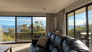 Ein modernes Wohnzimmer mit Ledersofa, Couchtisch und Blick auf einen See und Berge durch große Schiebeglastüren.