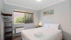 Ein Hotelzimmer mit Bett, Fenster mit Jalousien, Heizkörper, einer Kleiderstange, einem Nachttisch mit Lampe und einem Bild an der Wand.