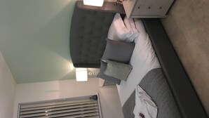 Ein Schlafzimmer mit Bett, Nachttisch, Lampe, Kommode und Fenster mit Jalousien.