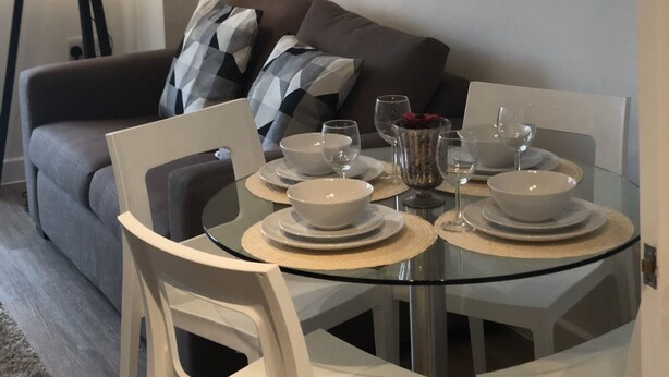 Ein Essbereich mit einem Glastisch, weißen Stühlen und einer grauen Couch mit gemusterten Kissen.