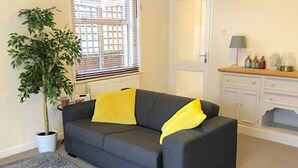 A grey sofa with yellow cushions, a potted plant, a grey rug, a white door, and a wooden cabinet with various items on top.