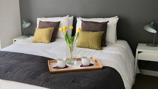 A neatly made bed with a tray of two cups and a vase of flowers, white bedding, and two bedside lamps.