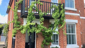 A two-story brick house with a balcony covered in ivy, a black door, and a small garden area.