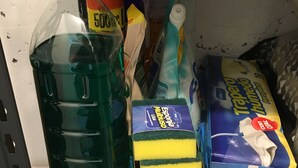 Een plank met schoonmaakmiddelen, waaronder een grote groene fles, gele en blauwe dozen en een rode bak.